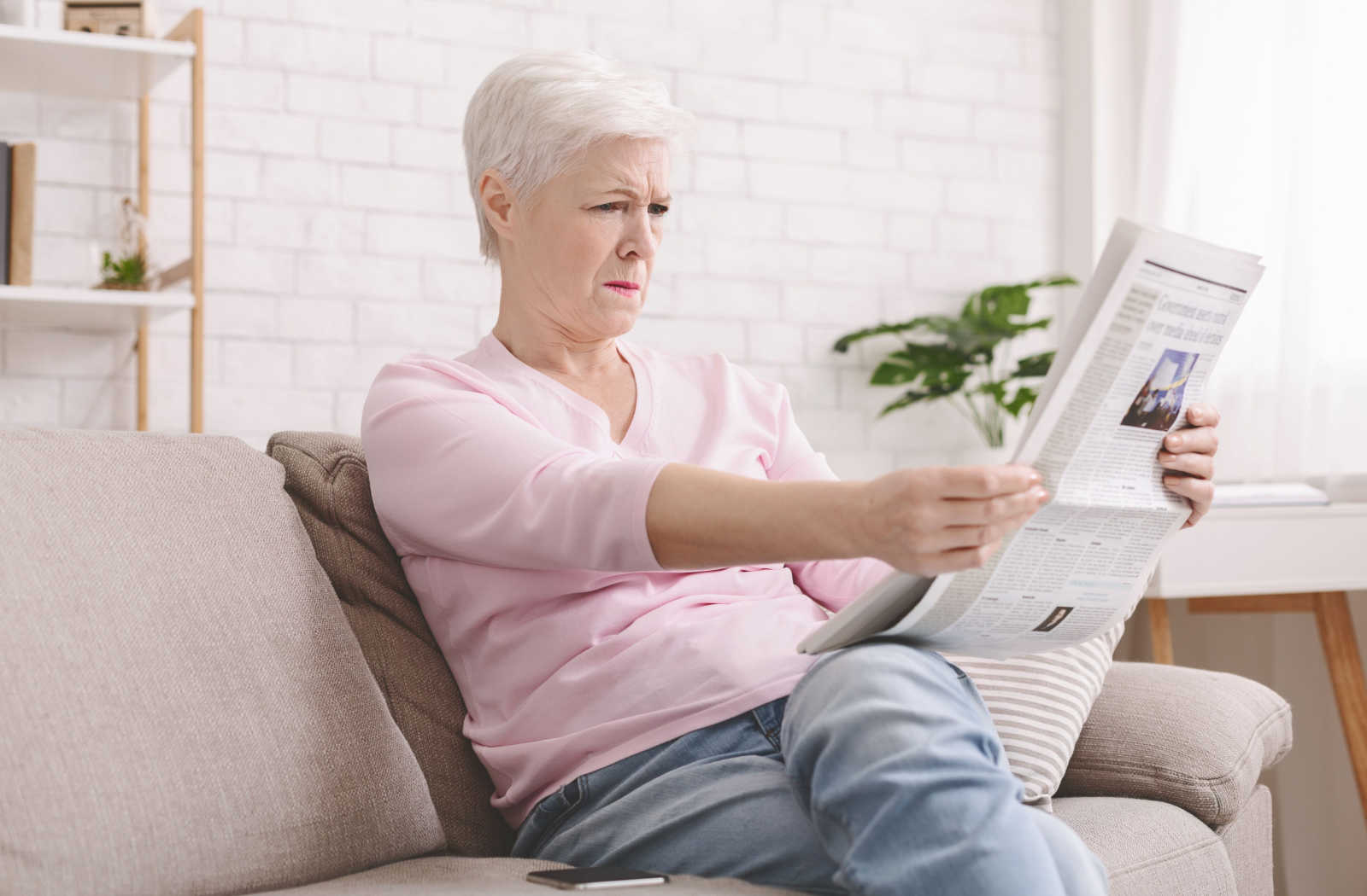 A woman in her fifties squinting her eyes as she has trouble reading the newspaper in her hand.