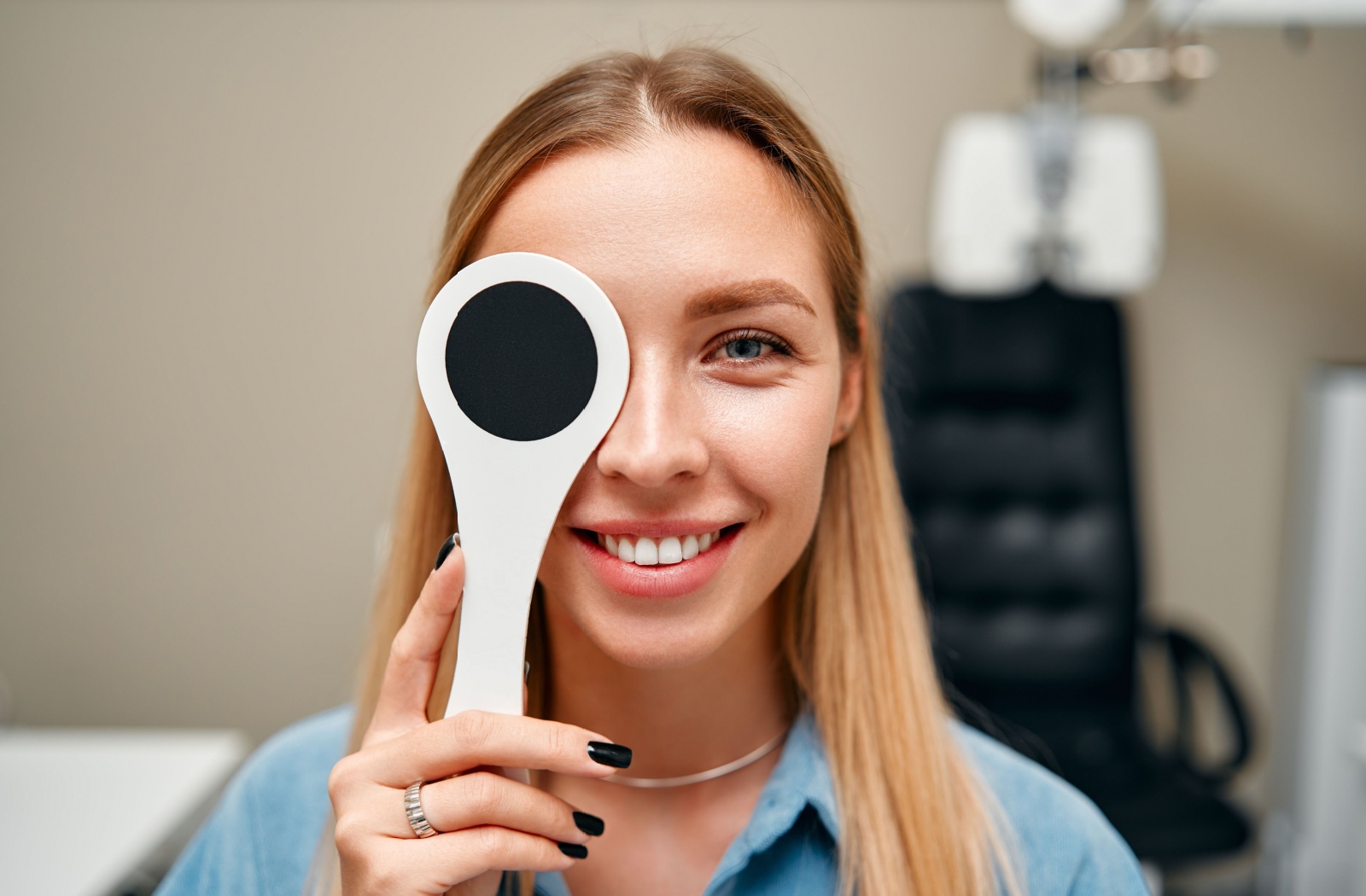 A patient in a blue shirt with nicely manicured fingernails smiles while holding a paddle that covers their right eye to test their vision while at the optometrist's office.