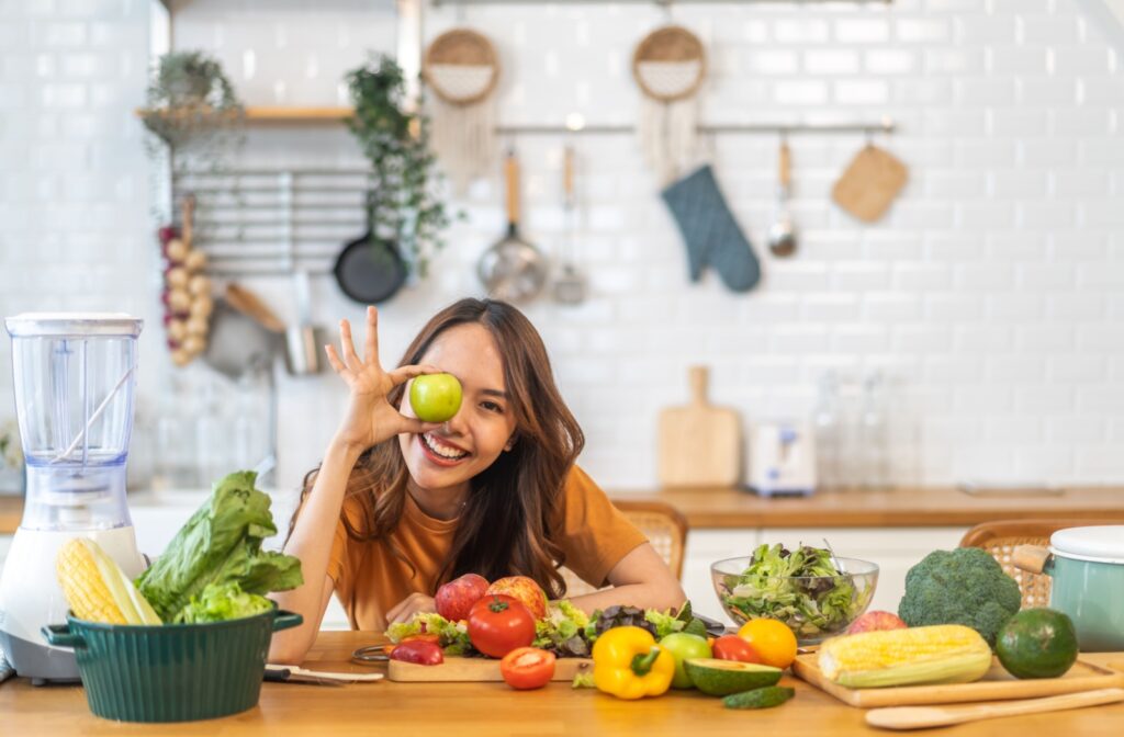 a woman holding up an apple against her eye with fruits and vegetables on her kitchen counter