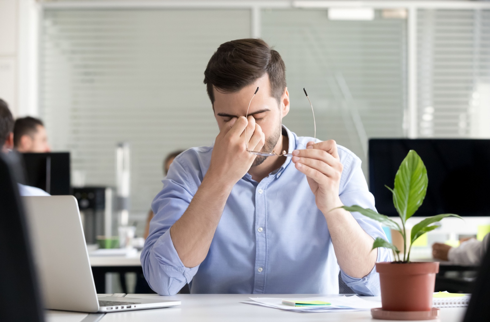 A young adult at work in an office, taking off their glasses and rubbing their eyes at their screen due to dry eyes.