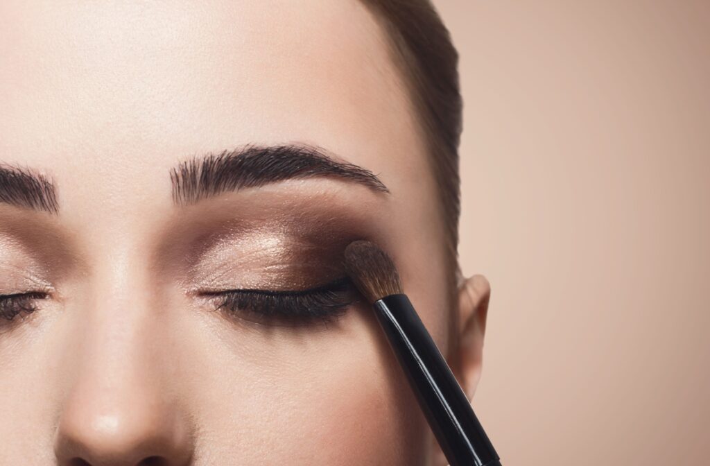 A black eyeshadow brush applied brown eyeshadow to a person's closed eyelid in an extreme close-up.