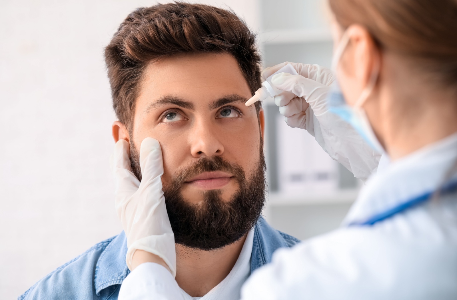 A patient looks upward as a healthcare provider in gloves and a face mask administers eye drops during an eye exam.