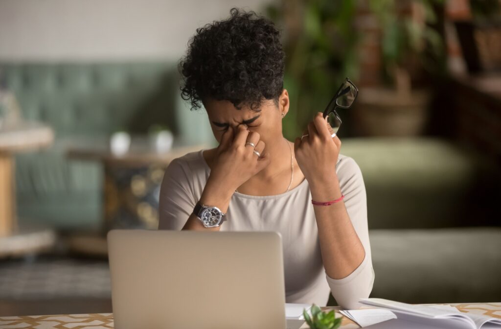 A person sits at a desk in front of a laptop, holding their glasses in one hand while rubbing their eyes with the other, showing signs of blurry vision.