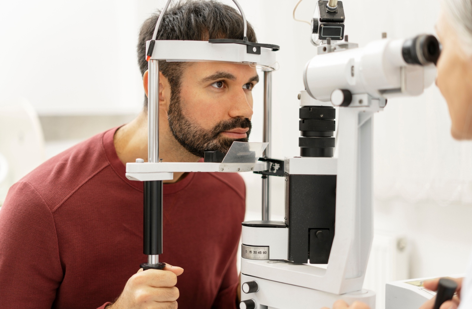 A man undergoes an eye exam using a slit lamp while an optometrist observes his eye closely.
