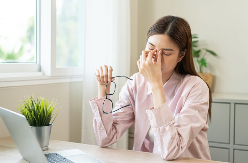 Woman rubbing her eyes while working at a computer, showing common symptoms of dry eye and digital eye strain.