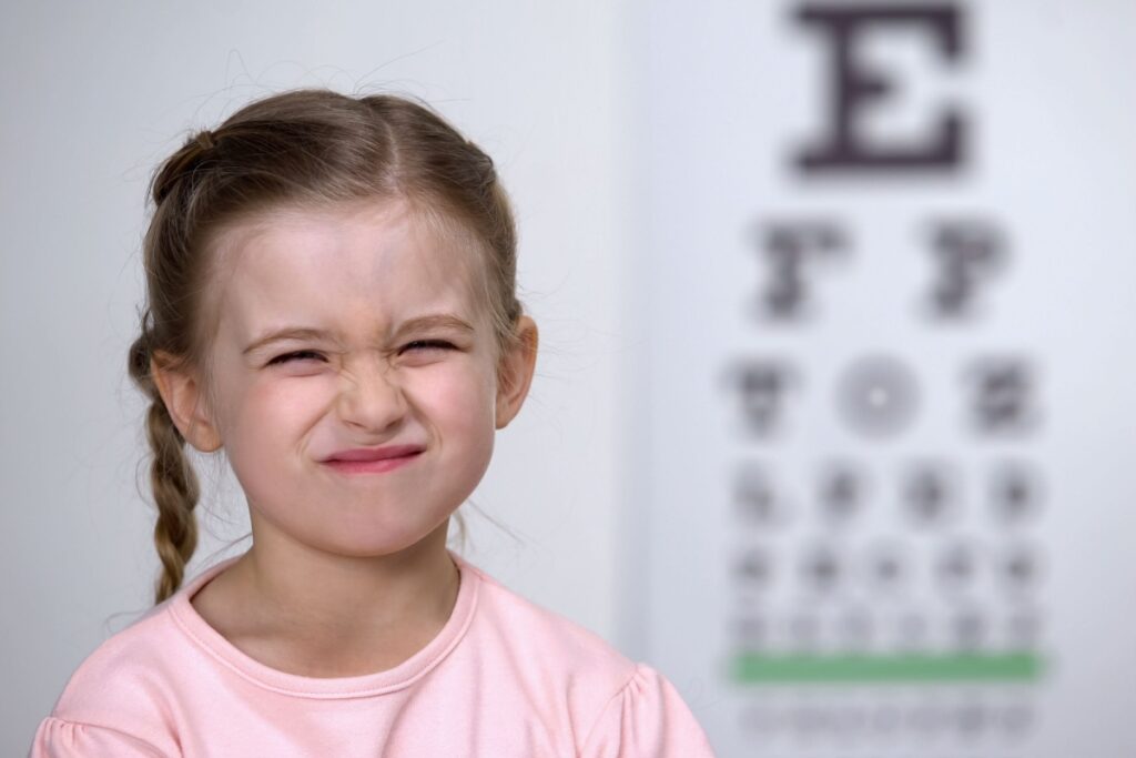 Young girl in pink shirt squinting during eye examination with blurred vision chart visible in background.