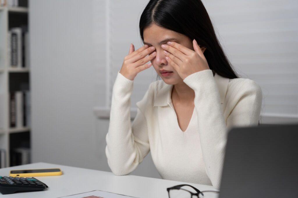 Young woman sitting at her desk rubbing her eyes due to dry eye discomfort.