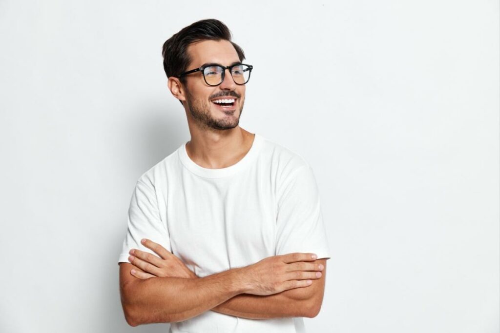 Attractive smiling man wearing glasses and standing crossed arm against a white wall.