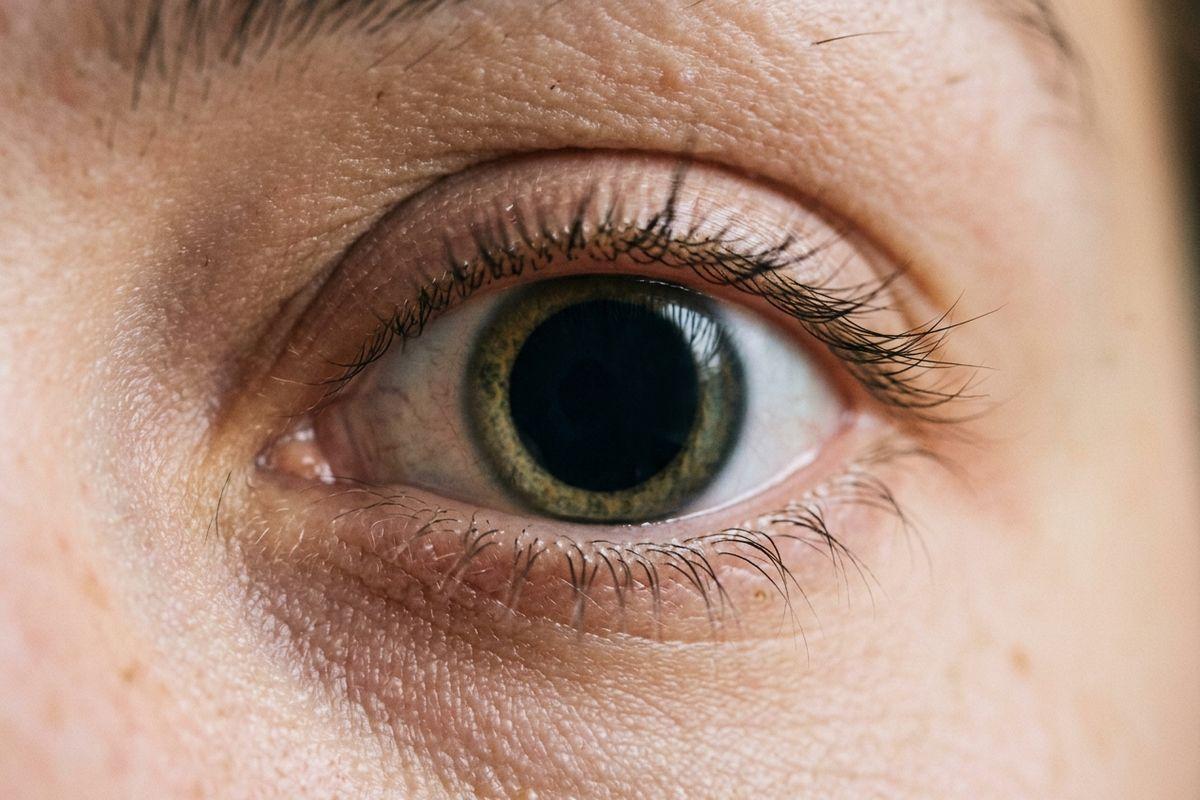 Extreme close-up of a person's open eye with a large black pupil, green-brown iris, dark lashes, and detailed skin texture around the eyelids.